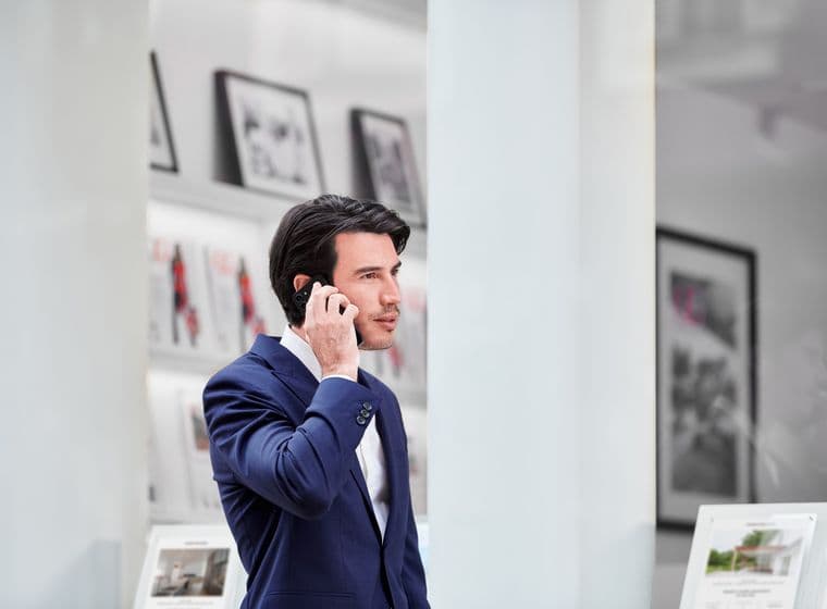 Man in a blue suit talking on a smartphone, standing in a modern office with framed pictures and brochures in the background.