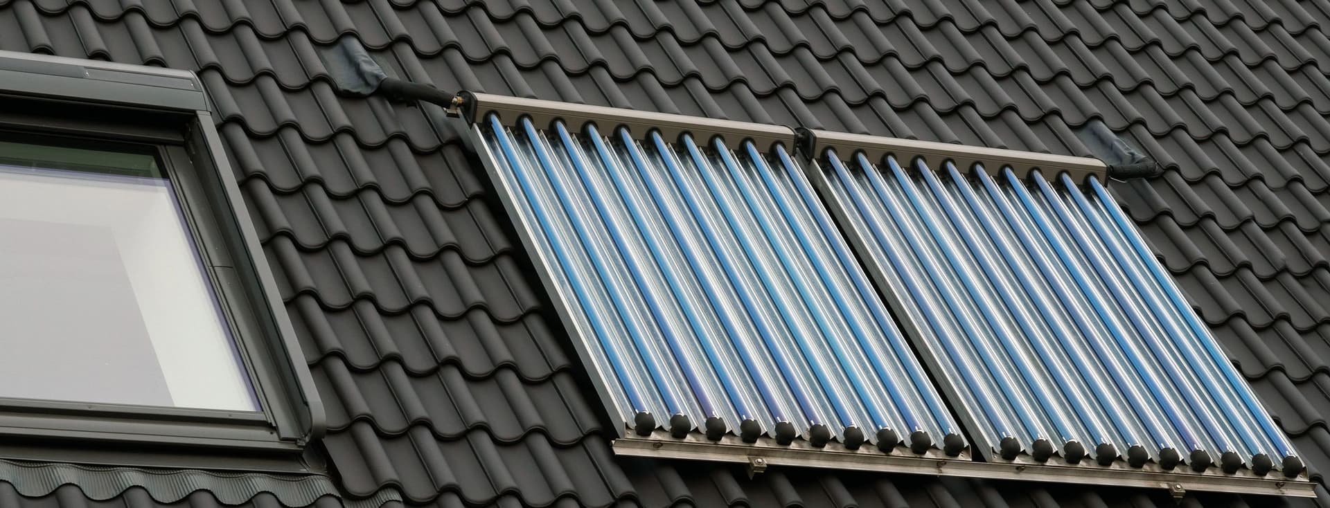 Two parallel rows of vacuum tube solar thermal collectors installed on dark gray roof tiles. A skylight window is visible on the left side of the collectors.