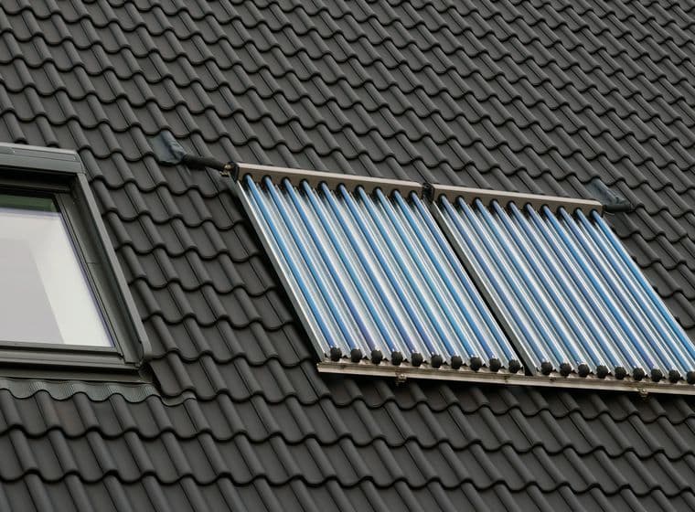 Two parallel rows of vacuum tube solar thermal collectors installed on dark gray roof tiles. A skylight window is visible on the left side of the collectors.