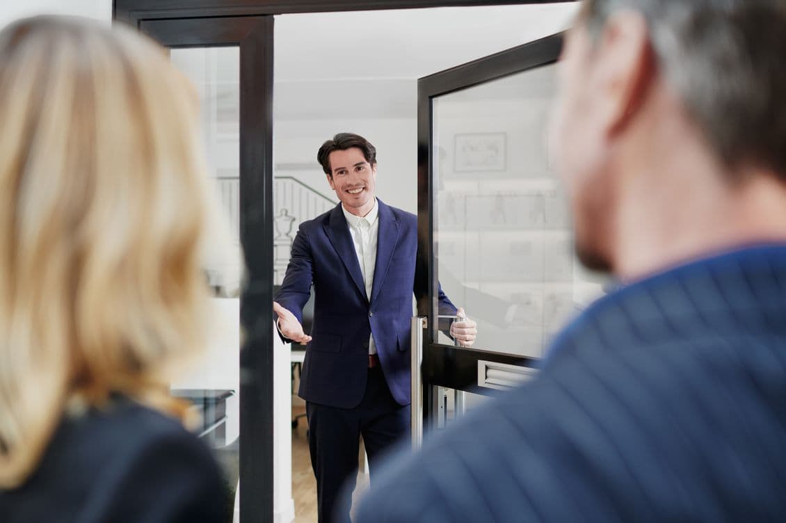 A man in a suit welcomes two people at a doorway, smiling and gesturing inside. The interior is modern and bright.