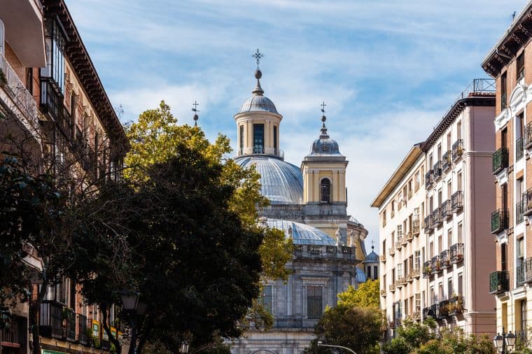 Vista de la Real Basílica de San Francisco el Grande en Madrid, España, enmarcada por edificios y árboles contra un cielo azul.