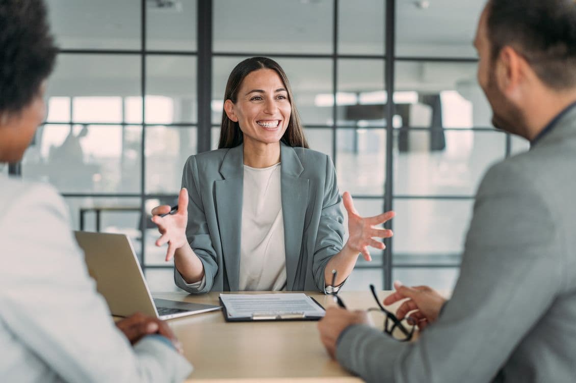 Smiling woman in a gray blazer gestures while meeting with two clients at a table with a laptop and paperwork.