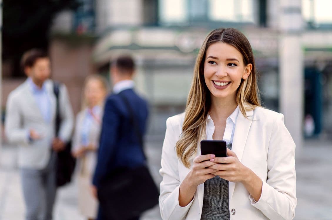 A smiling woman in a white blazer holds a smartphone outdoors.
