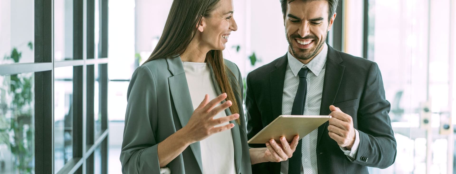 Um homem e uma mulher sorrindo, vestidos com ternos de negócios, olham para um tablet em um escritório moderno.