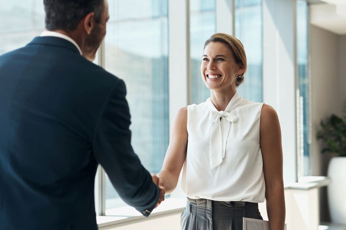 A smiling woman in a white blouse shakes hands with a man in a suit in a modern office.