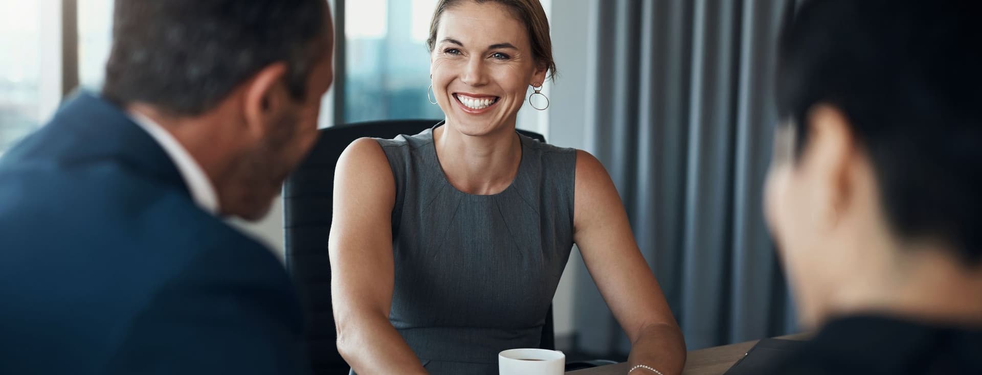 Uma mulher sorridente em um vestido cinza está sentada a uma mesa com dois homens de terno.