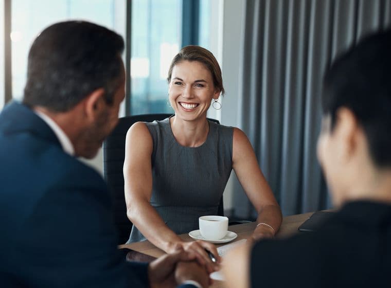 Uma mulher sorridente em um vestido cinza está sentada a uma mesa com dois homens de terno.