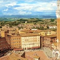 Luftaufnahme von Siena, Italien, mit Blick auf die Piazza del Campo mit ihren charakteristischen roten Dächern und dem hohen Torre del Mangia unter einem bewölkten Himmel.