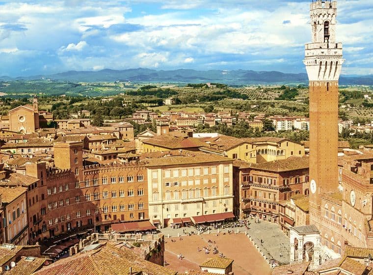 Luftaufnahme von Siena, Italien, mit Blick auf die Piazza del Campo mit ihren charakteristischen roten Dächern und dem hohen Torre del Mangia unter einem bewölkten Himmel.