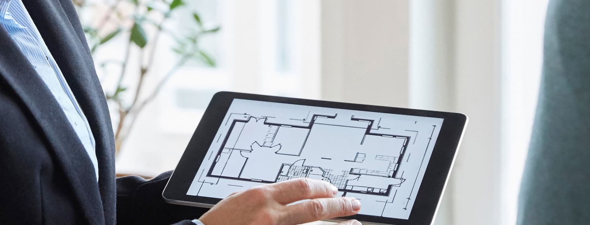 Ein Agent im Anzug hält ein Tablet, auf dem ein Grundriss eines Hauses angezeigt wird.