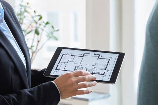 Real estate professional in business suit holding tablet displaying architectural floor plan, photographed in bright modern interior with window and plant in background