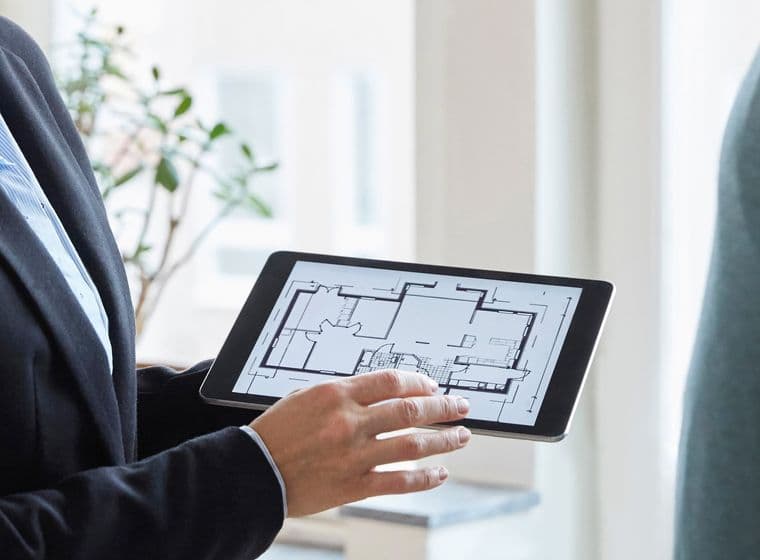 Ein Agent im Anzug hält ein Tablet, auf dem ein Grundriss eines Hauses angezeigt wird.