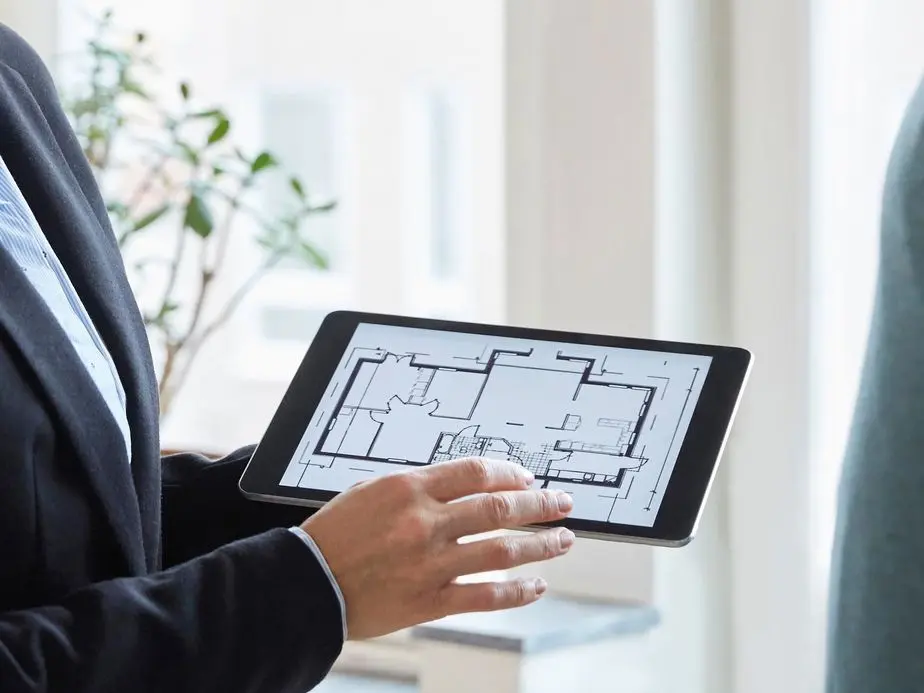 Agent in suit holds a tablet displaying a home floor plan.