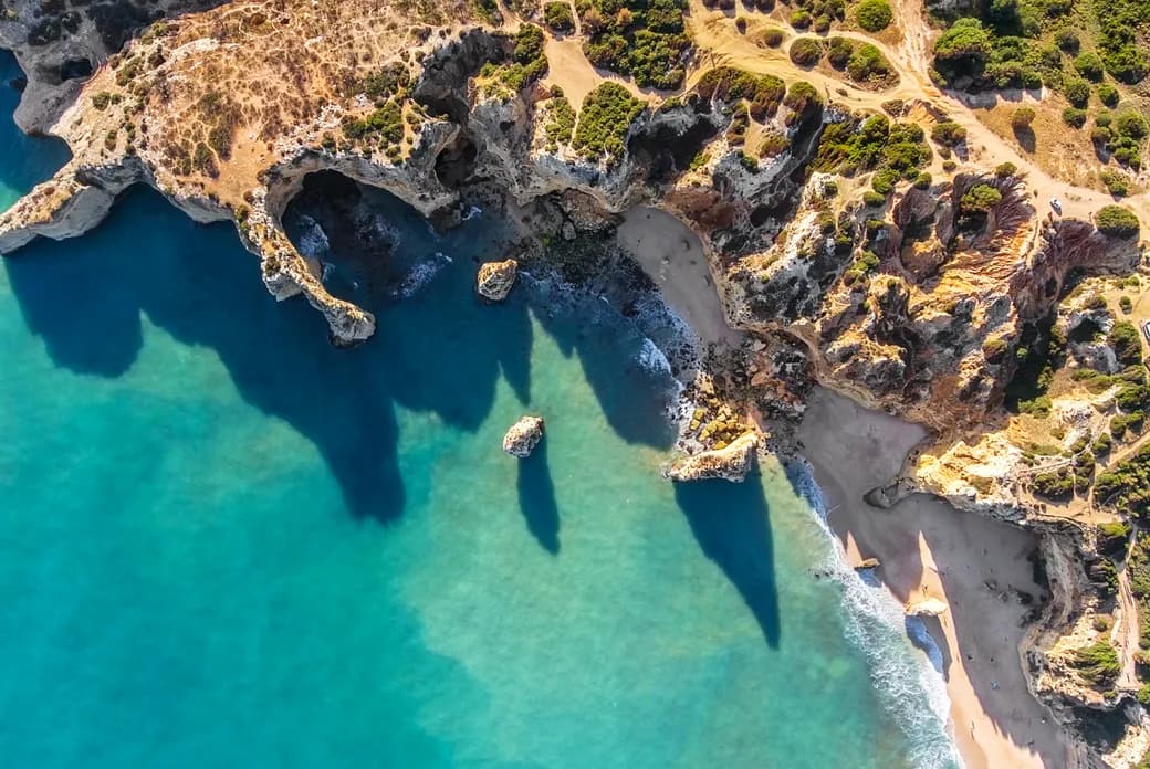 Aerial view of rugged coastline with turquoise water, rocky cliffs, a sandy beach, and green hills with winding dirt roads.