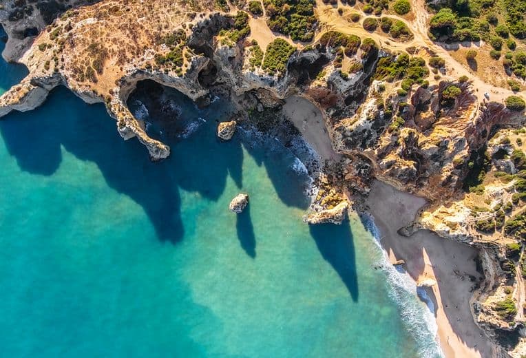 Aerial view of rugged coastline with turquoise water, rocky cliffs, a sandy beach, and green hills with winding dirt roads.