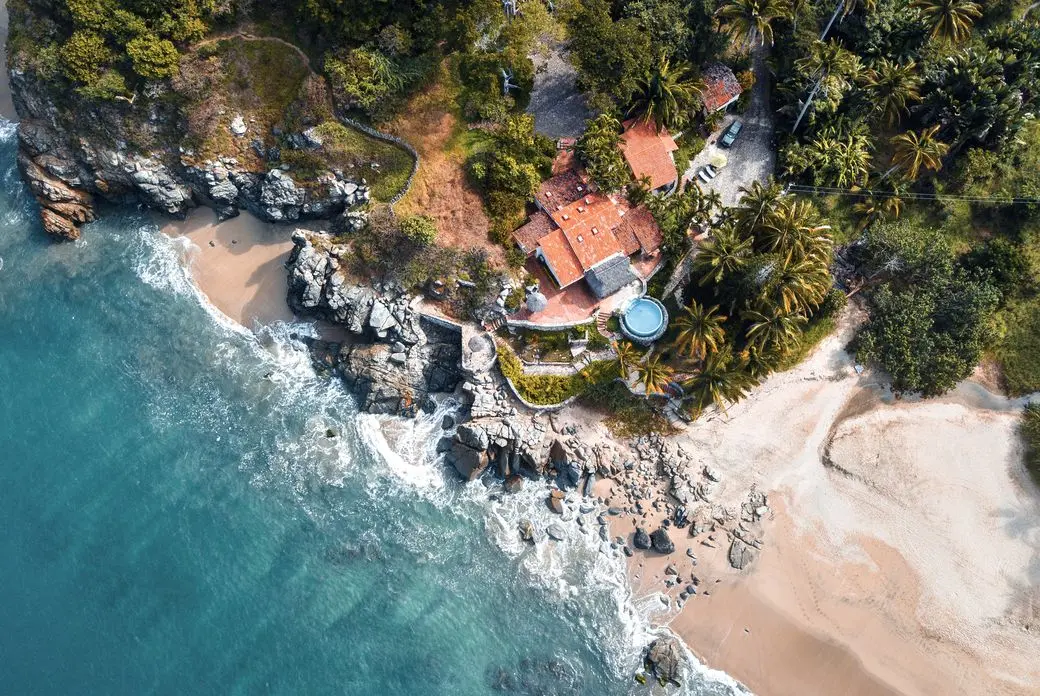 Aerial view of a tropical beach with turquoise water, waves, and a villa with a red tile roof surrounded by lush green trees.