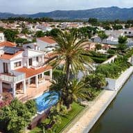 Drone view of one of Empuriabrava's canals with a modern white house with a swimming pool and the urbanisation and mountain views on the second plan