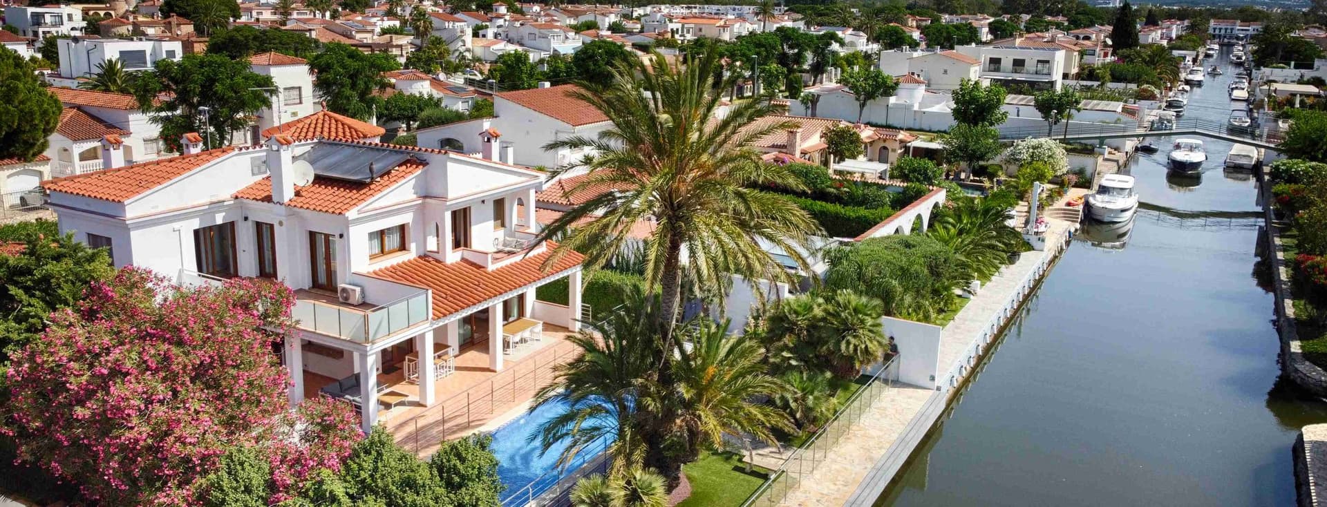 Drone view of one of Empuriabrava's canals with a modern white house with a swimming pool and the urbanisation and mountain views on the second plan