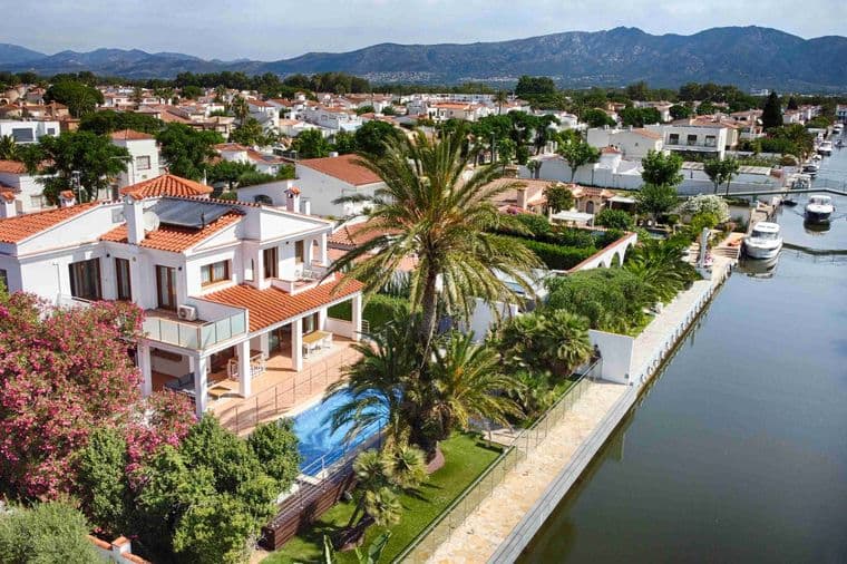 Drone view of one of Empuriabrava's canals with a modern white house with a swimming pool and the urbanisation and mountain views on the second plan