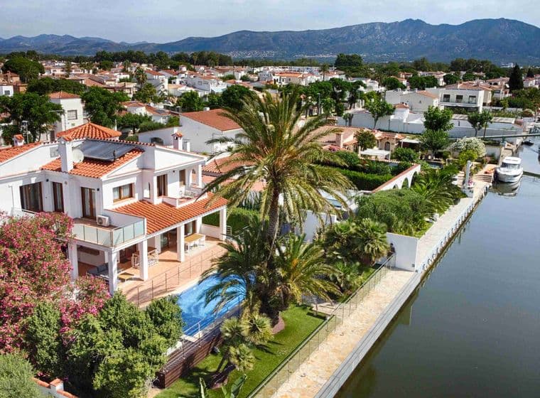 Drone view of one of Empuriabrava's canals with a modern white house with a swimming pool and the urbanisation and mountain views on the second plan