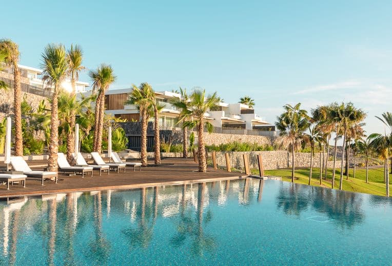 Infinity pool at a tropical resort with palm-lined wooden deck, white modern villas, and a clear blue sky.