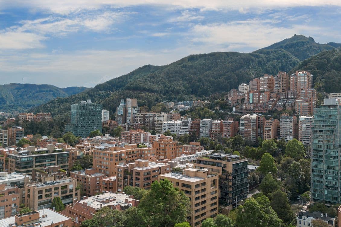 Vista panorámica de Bogotá, Colombia, con edificios de ladrillo, árboles verdes y montañas bajo un cielo azul con nubes blancas.