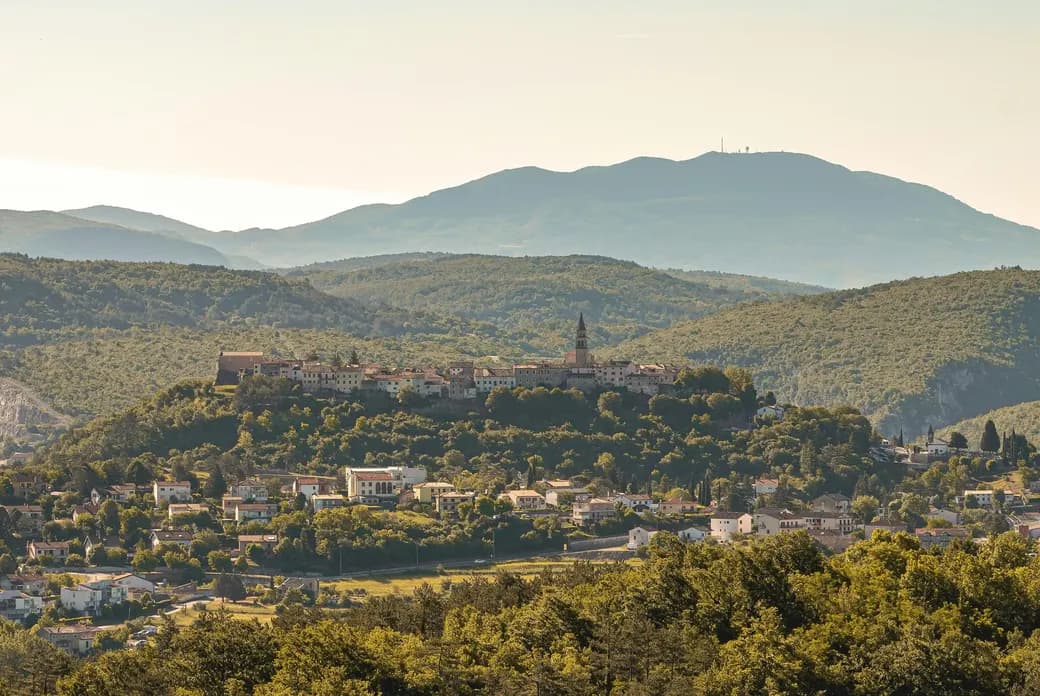 Panoramic view of a mountain village in Istria with a church tower, surrounded by green hills and mountains in the background, captured in warm evening light