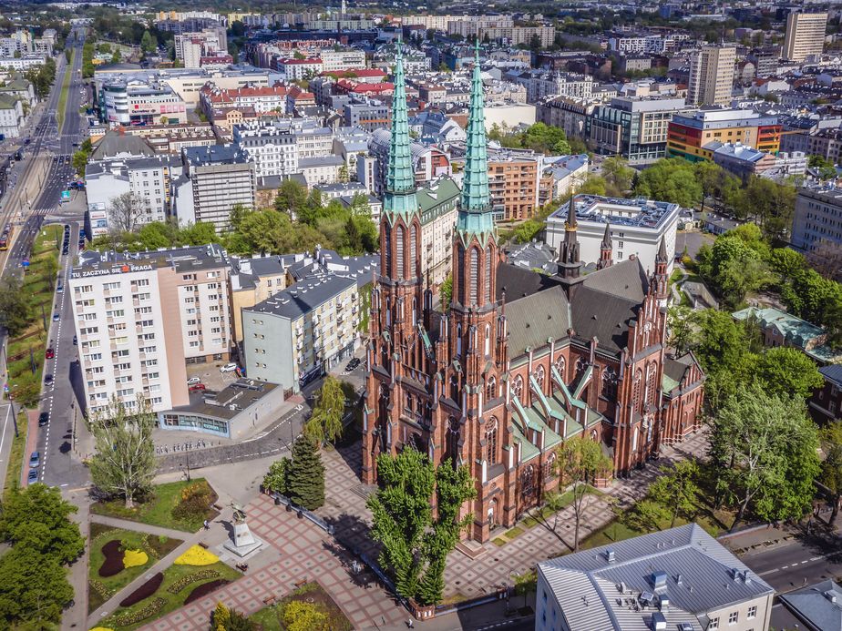 Aerial view of a red brick cathedral with two tall spires, surrounded by trees and urban buildings in a bustling city.