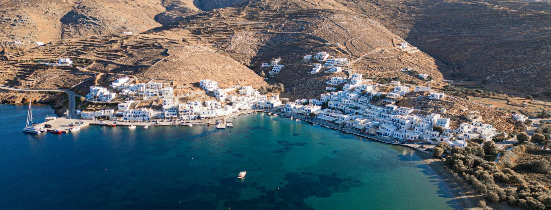 Aerial view of a coastal Greek village with white houses along a turquoise bay, small boats, and dry brown hills behind.