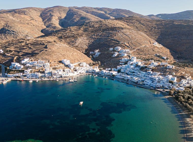 Aerial view of a coastal Greek village with white houses along a turquoise bay, small boats, and dry brown hills behind.