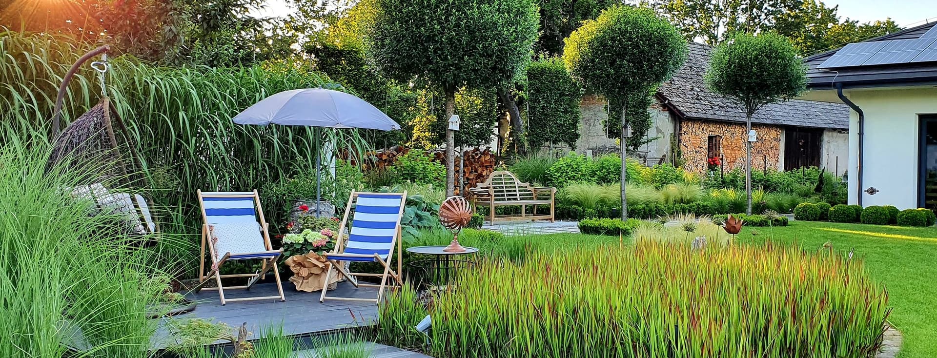 Ein üppiger Garten mit gestreiften Stühlen, einem blauen Sonnenschirm und einer Korbschaukel. Das Sonnenlicht fällt durch die Bäume und lässt das satte Grün strahlen