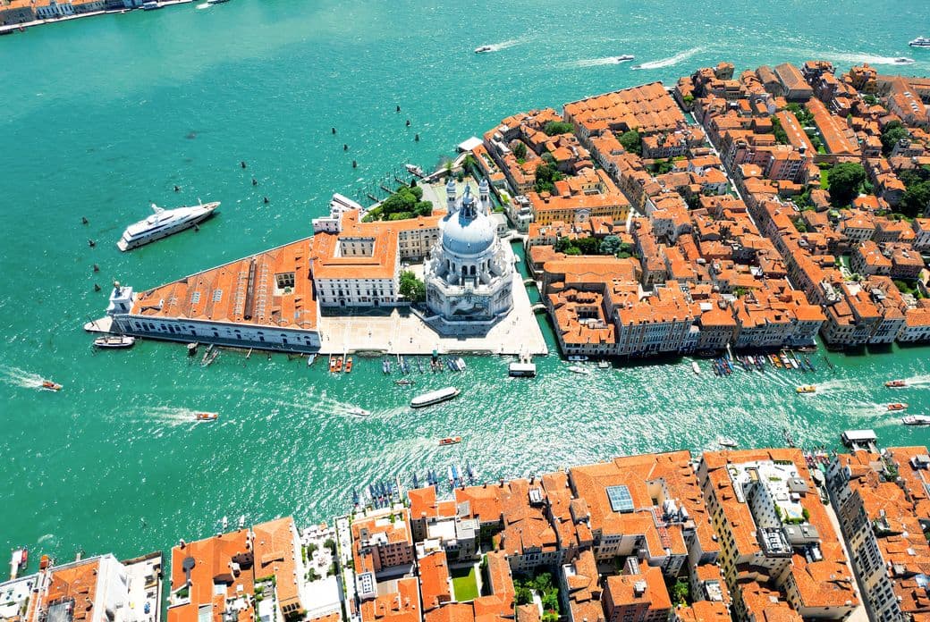Aerial view of Venice, featuring turquoise canals, red-tiled rooftops, and a prominent white-domed building near the water.