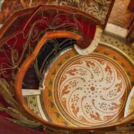 Ornate spiral staircase with red carpet and decorative wrought-iron railing, set on a colorful mosaic floor and marble walls.