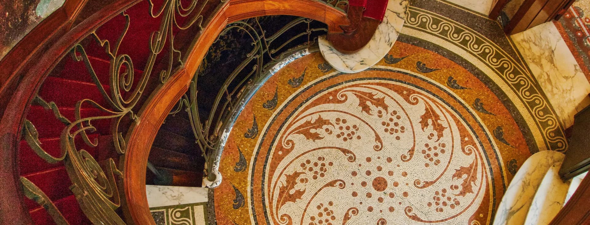Ornate spiral staircase with red carpet and decorative wrought-iron railing, set on a colorful mosaic floor and marble walls.