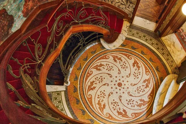 Ornate spiral staircase with red carpet and decorative wrought-iron railing, set on a colorful mosaic floor and marble walls.