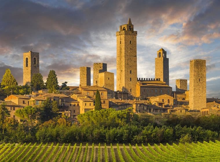 Mittelalterliche Stadt mit zahlreichen Steinturmen, umgeben von üppigen Weinbergen und Grünflächen, unter einem dramatischen, bewölkten Himmel bei Sonnenuntergang.
