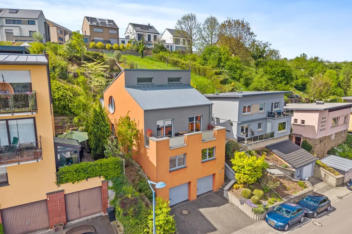 Hillside neighborhood of modern pastel houses surrounded by greenery, with parked cars along a curved street and an orange building in the foreground.
