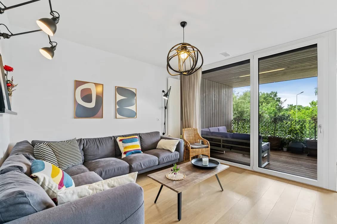 Bright living room with a gray sectional, colorful cushions, abstract wall art, an orb pendant light, and sliding glass doors to a sunny balcony.