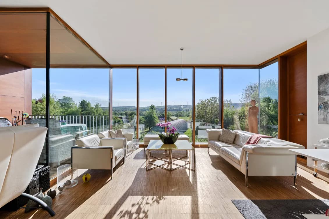 Bright modern living room with expansive glass walls, white sofas, and a glass coffee table, overlooking a landscape with trees and wind turbines.