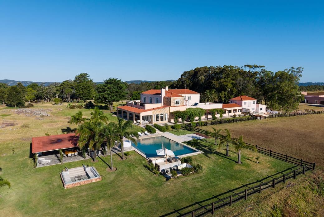 Aerial view of a countryside estate: pink villa with red tile roofs, swimming pool, palm trees and fenced lawns under a clear blue sky.