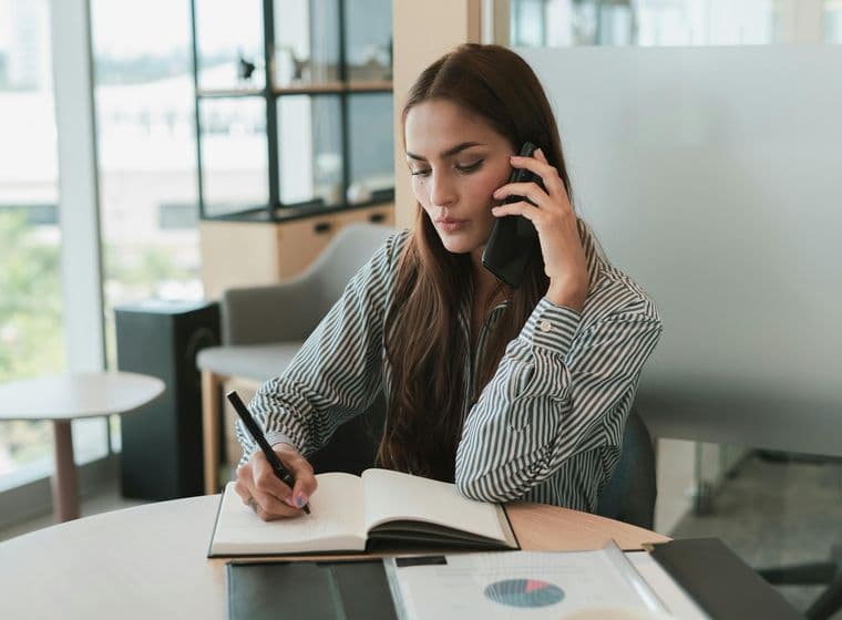 Eine Frau in einem gestreiften Hemd sitzt an einem runden Tisch, telefoniert und schreibt in ein Notizbuch.