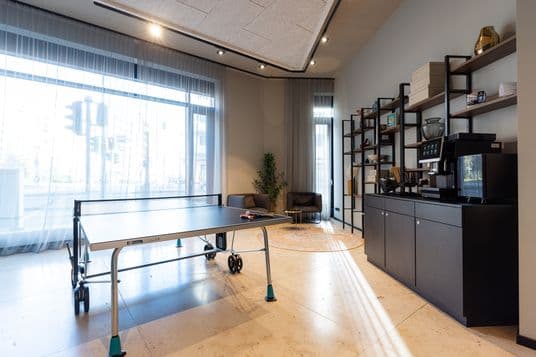 Modern lounge with a ping-pong table in the foreground, large windows with sheer curtains, and a tall black shelving unit filled with boxes and decor.
