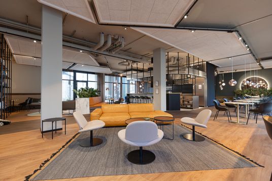 Contemporary open-plan lounge with an orange sofa, gray chairs, round tables, hanging pendant lights, exposed ceiling pipes, and lush plants.