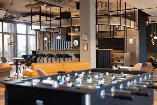 Modern cafe interior with glass partition, warm pendant lighting, marble bar, black stools, large windows, and a foosball table in the foreground.
