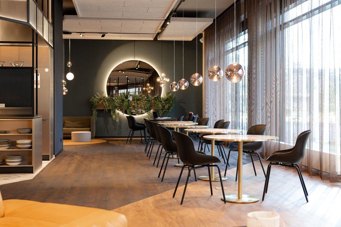 Modern cafe with round marble tables, dark chairs, pendant glass globe lights, and large windows with sheer curtains and a plant wall.