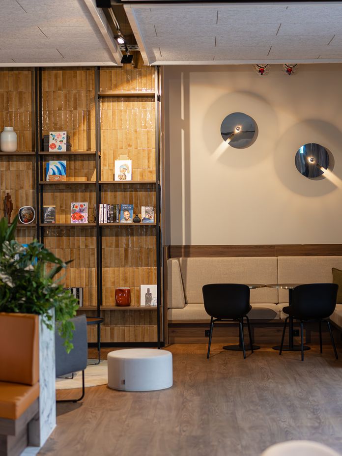 Modern lounge with tall wooden shelves, tan tile wall, lush plants, and a beige seating nook with two round mirrors by large windows.