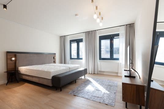 Modern bedroom with an upholstered bed, wooden headboard, small round side table, cluster pendant lights, large windows with sheer gray curtains, and a TV on a wooden stand.