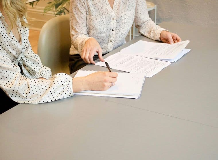 se ven dos mujeres con camisa blanca con topos negros, firmando una escritura