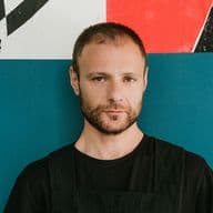 Ένας άντρας με γένια στέκεται μπροστά σε έναν μπλε τοίχο με αφίσες.