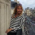 Smiling woman in a black-and-white striped blouse leans on a balcony railing overlooking a busy city street with ornate buildings.